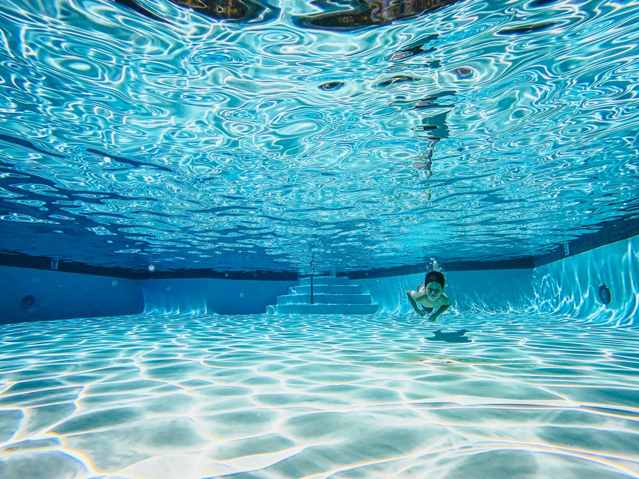 gallery-6 A young child swimming underwater in a clear blue pool, showcasing playful aquatic motions.
